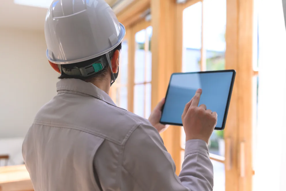 ヘルメット姿でタブレットを見ながら住宅を診断する調査員